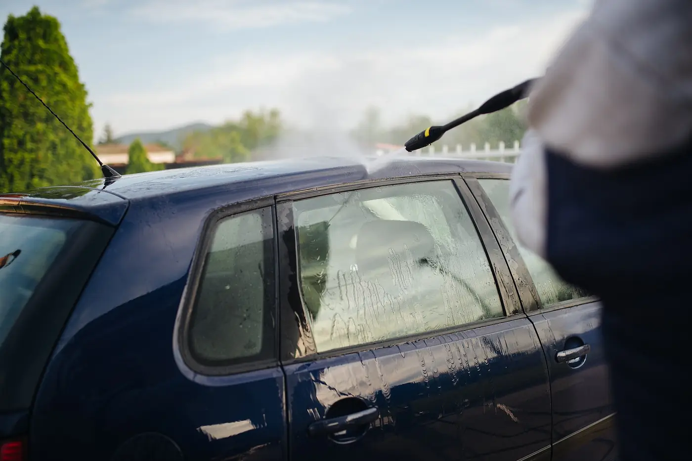 Person pressure-washing a dark blue hatchback on a driveway