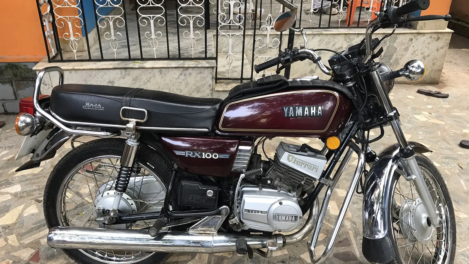Maroon Yamaha RX100 parked in front of a house with a decorative iron gate