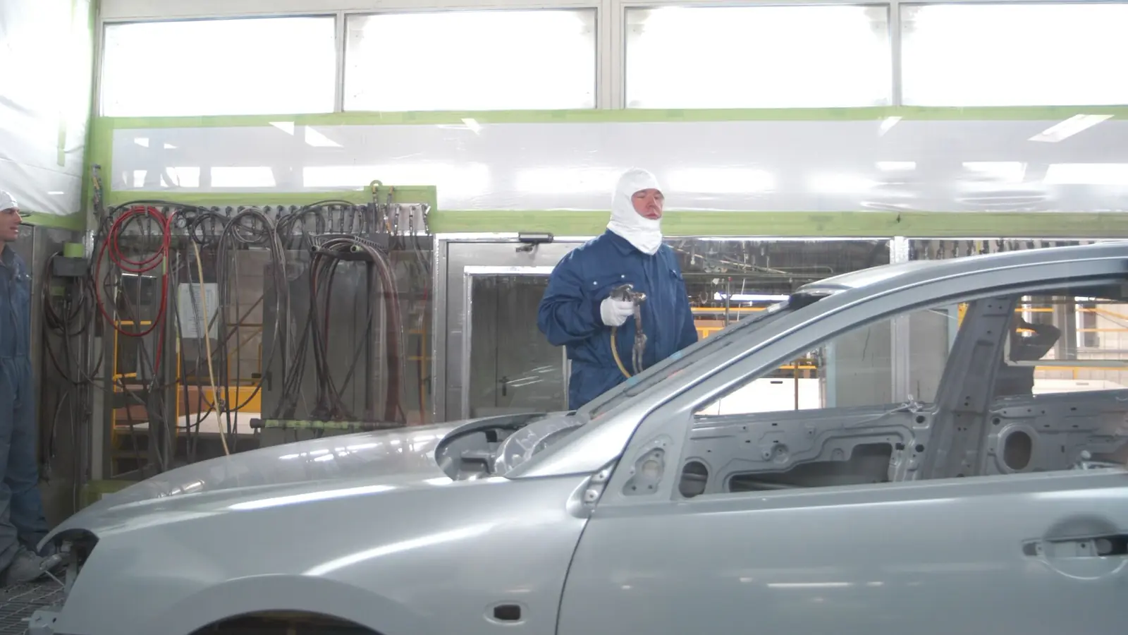Painter in protective overalls and hood spraying a silver car inside a spray booth