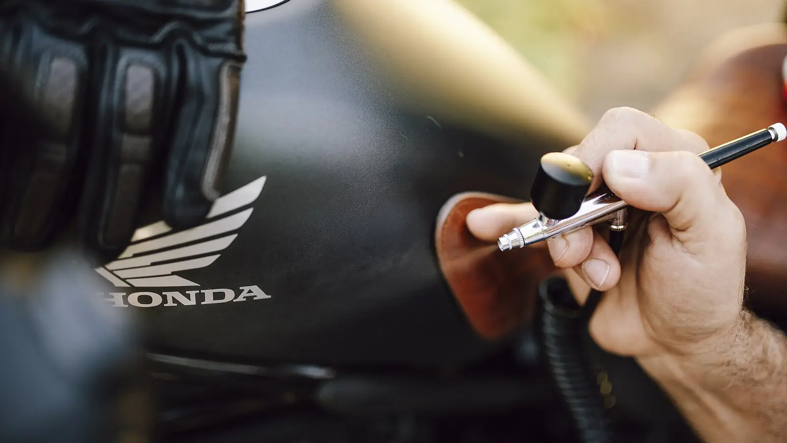 Close-up of an artist's hand airbrushing a design onto a black motorcycle fuel tank