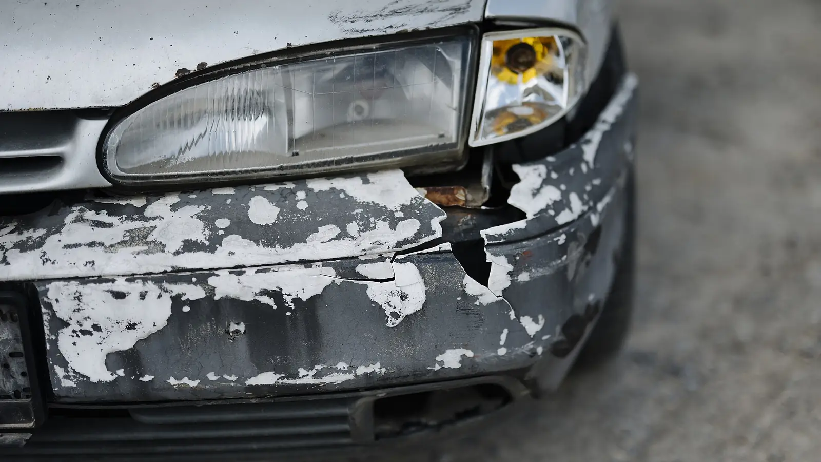 Close-up of a silver car's front corner with paint flaking off around the headlight and bumper
