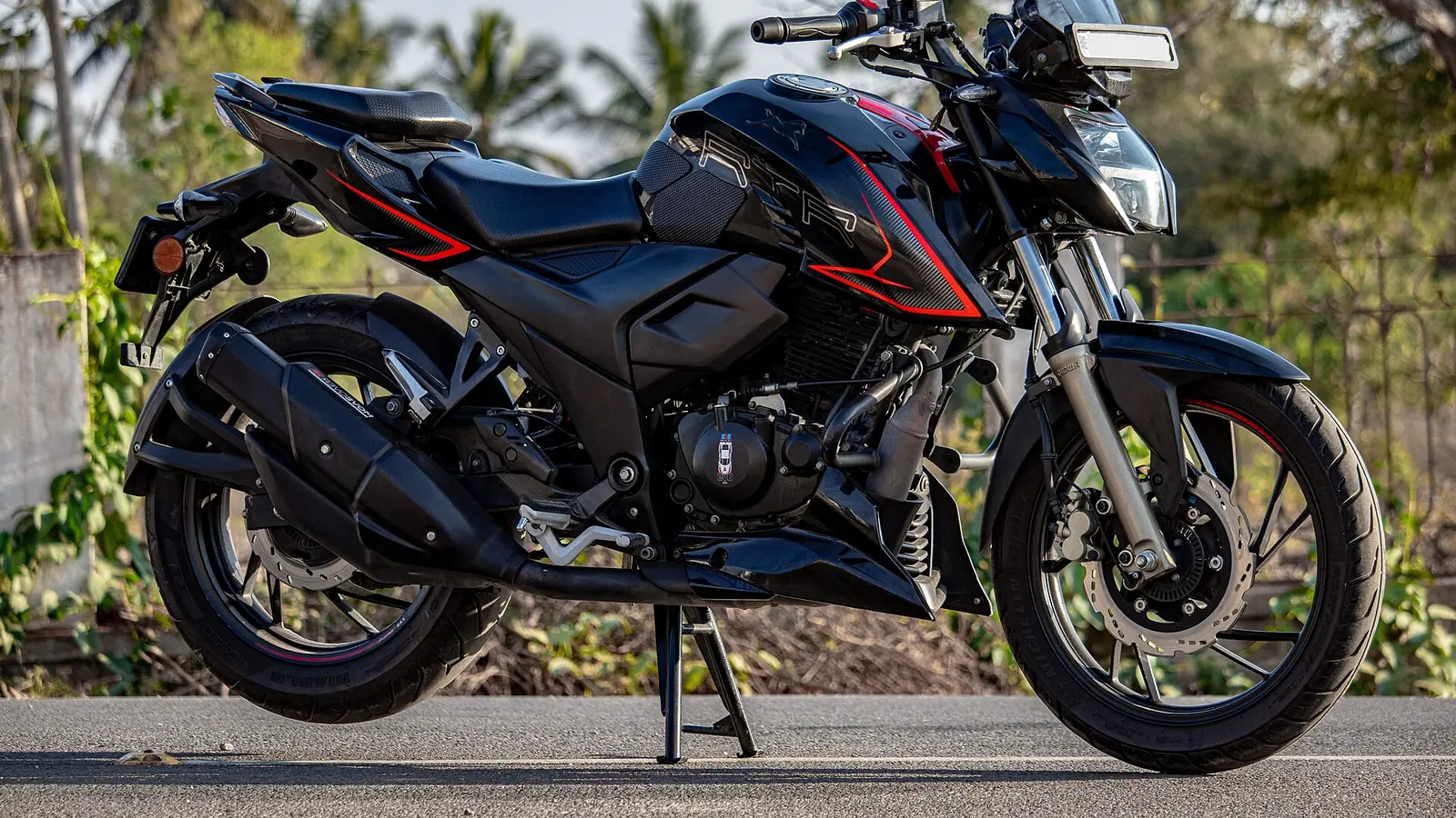 Black TVS Apache RTR 200 parked on a road with palm trees behind it